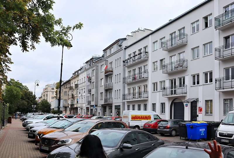 Ulica zastawiona po prawej i lewej samochodami. Z lewej zielone drzewa liściaste. Z prawej kamienica przy kamienicy ciągną się niemal po horyzont.