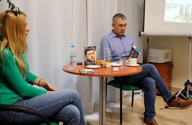 Krótko przystrzyżony mężczyzna w średnim wieku siedzi przy stoliku. Na stoliku leży filiżanka, woda w butelkach i dwie książki. Po prawej, obok mężczyzny, stoi ekran rzutnika, a na nim zdjęcie bloków mieszkalnych na tle dymiących kominów fabrycznych. Z lewej strony siedzi młoda blond kobieta w dżinsach i zielonym swetrze.