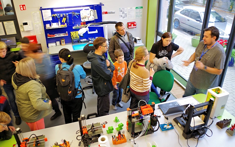 Kilkanaście osób stoi w pracowni technicznej, słuchając informacji od pracownika placówki. Na stole znajdują się cztery drukarki 3 d. Po prawej stronie przeszklone okno z widokiem na parking. W tle ściana z narzędziami do majsterkowania.