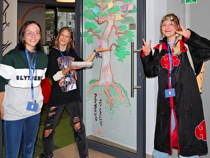Trzy uśmiechnięte nastolatki. Jedna ubrana w stylu postaci z komiksów mangi. Druga wskazuje szklane malunek na szklanych drzwiach. Przedstawia on drzewo życzeń, na którym przyklejonych jest kilka kartek z życzeniami.