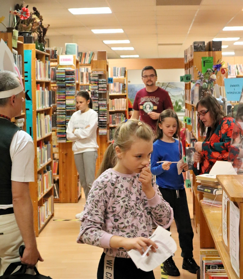Oświetlone pomieszczenie biblioteczne. Kilka osób, większość dzieci spoglądających na książki. Z prawej pracownik placówki wyjaśnia coś dziewczynce.