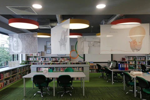 Widok na salę biblioteczną, pod sufitem zawieszone plansze wystawy z ilustracjami z książki „Co robią uczucia?”.