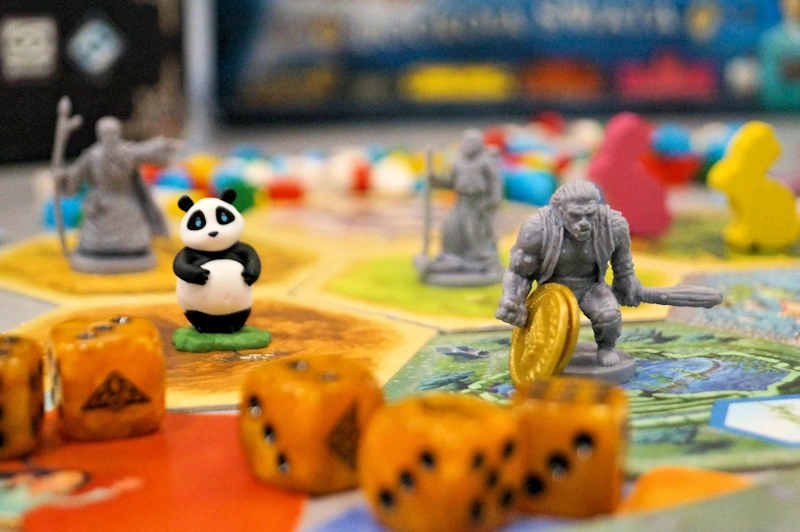 Gra planszowa widoczna z bliska. Na niej porozsypane są kości oraz figurki różnych fantazyjnych postaci oraz pandy.