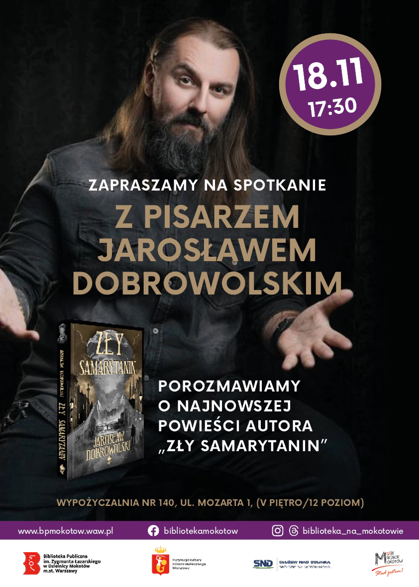 Mężczyzna w długich ciemnych włosach oraz dłuższą brodą i wąsami, ubrany na czarno patrzy się z uśmiechem w obiektyw, rozkładając ręce.