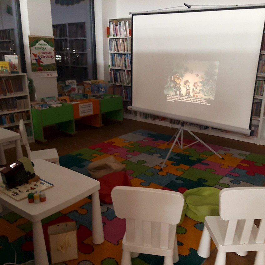 Kolorowy, łaciaty dywan, na którego końcu stoi biały ekran z wyświetlaną na nim animowaną bajką wyświetlaną ze starego projektora domowego.