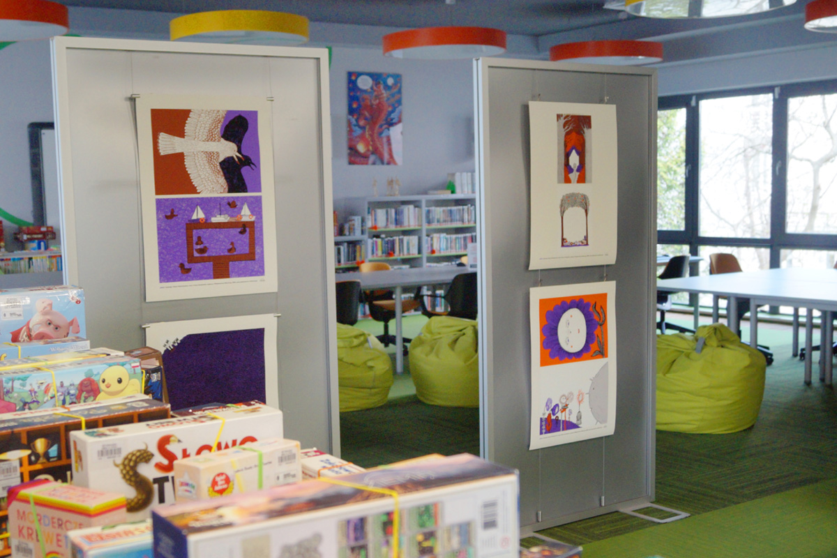 Zdjęcie przedstawia dwa ekspozytory z czterema kolorowymi ilustracjami w przestrzeni sali bibliotecznej.