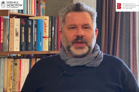 Mężczyzna w średnim wieku, z siwiejącymi włosami, brodą oraz wąsami patrzy się wprost do kamery. Ubrany w granatowy sweter i apaszkę. Za nim po lewej półki z książkami. Po prawej bordowo-szara zasłona okienna.