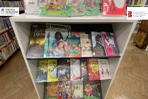 Biała szafka a w niej i na niej przykładowe propozycje komiksów i książek dostępnych w bibliotece dla dzieci i młodzieży numer trzydzieści trzy.