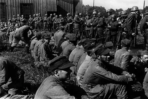 Żołnierze siedzący na ziemi, jeńcy z Westerplatte, pilnowani przez żołnierzy niemieckich z karabinami. 