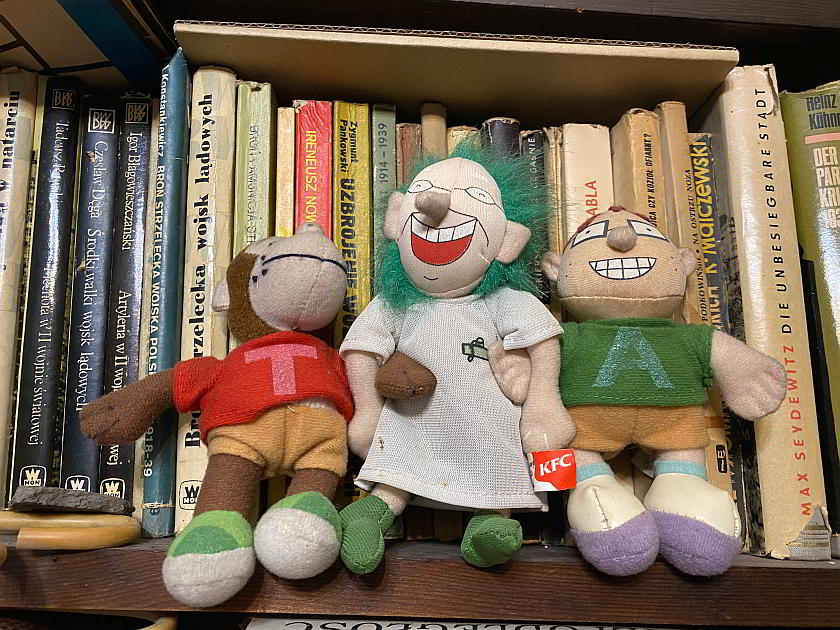 Na zdjęciu znajdują się trzy pluszowe zabawki przedstawiające kolejno: Tytusa, Profesora T’Alenta i A’Tomka. Tytus ubrany jest w czerwoną koszulkę z białą literą T. Profesor T’Alent ma na sobie okulary oraz biały kitel naukowca. Włosy profesora są zielone i mocno zmierzwione. Widoczny jest też jego szeroki uśmiech. A’Tomek ubrany jest w zieloną koszulkę z białą literą A. Na nosie ma okulary. Również szeroko się uśmiecha. Wszystkie pluszanki trzymają się za ręce. W tle znajduje się kolekcja najróżniejszych książek.