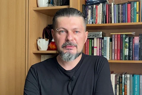 Pisarz ubrany w czarny podkoszulek patrzy prosto w kamerę. Ma krótką siwiejącą brodę oraz wąsy. Do prawej brwi ma przyczepiony kolczyk. W tle widać półki z książkami.