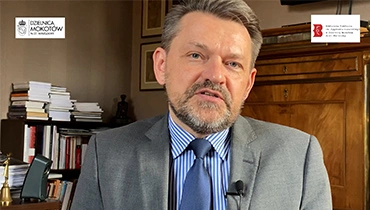 Mężczyzna w garniturze siedzi w pomieszczeniu na tle stylowej, brązowej komody i książek.