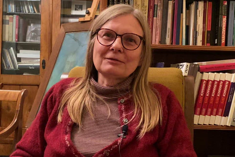 Blondwłosa pisarka w okularach siedzi na krześle i uśmiecha się do widza. Za nią stoją półki z książkami.