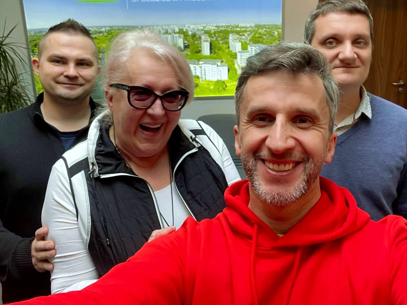 Trzech mężczyzn i kobieta uśmiechają się do obiektywu. Na przodzie redaktor radia warszawa w czerwonej bluzie. Za nim dyrektor biblioteki mokotowskiej w jasnym stroju i w ciemnych okularach. Z tyłu dwóch pracowników biblioteki w ciemnych bluzach.