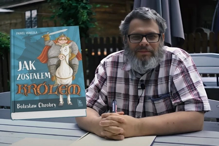 Mężczyzna z brodą siedzi przy stole, trzyma w ręku długopis, po lewej wmontowana okładka jego książki