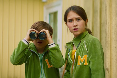 Kadr z filmu: dwójka przyjaciół dziewczynka i chłopiec patrzą przed siebie, chłopiec spogląda przez lornetkę. Na sobie mają zielone bluzy, ona z dużą literą „M”, on z „L”. W tle widać dom.