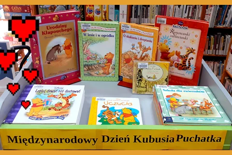 Stojak z na książki, na którym zostały umieszczone pozycje związane z Kubusiem Puchatkiem. Pod spodem tekst: międzynarodowy dzień Kubusia Puchatka. Z lewej kilka dorysowanych serduszek. W tle półka z książkami.