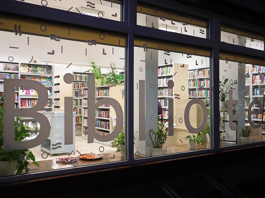 Zdjęcie frontu biblioteki dziecięcej, z naklejonym na szybach napisem biblioteka. Widać oświetlone wnętrze z licznymi regałami zapełnionymi książkami.