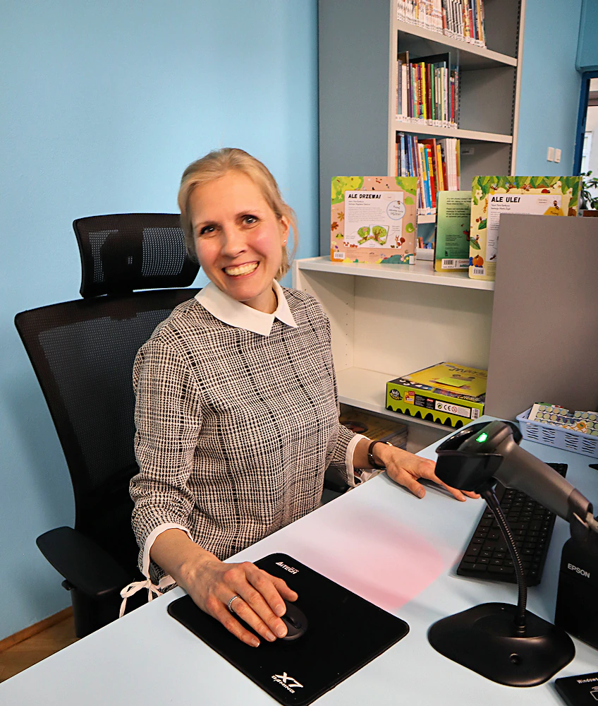 Bibliotekarka siedzi prze komputerem, szeroko uśmiechając się do obiektywu.