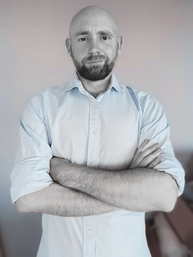 Zdjęcie łysego mężczyzny z brodą w jasnej koszuli wpatrzonego w obiektyw.