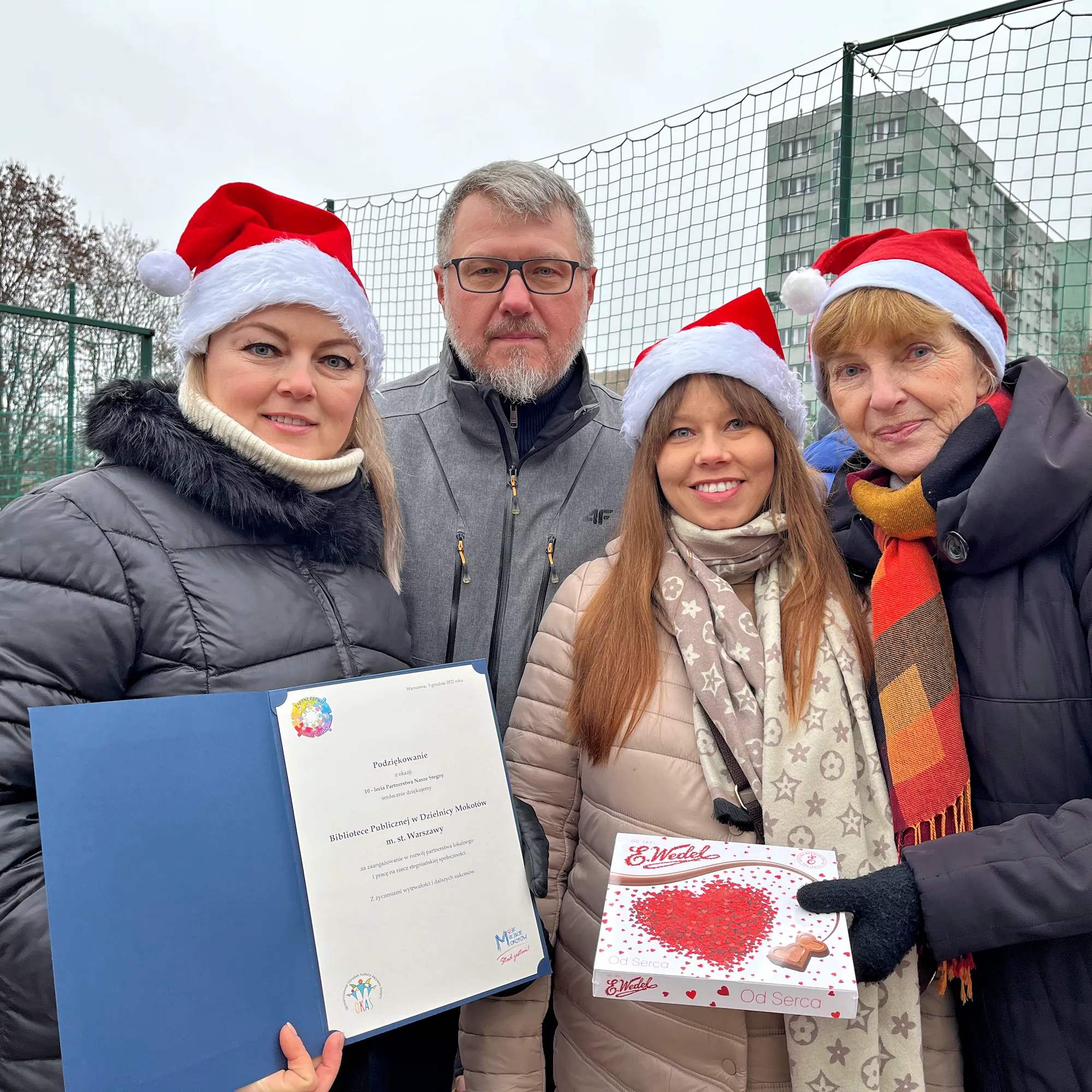 Zimowa aura. Trzy kobiety w zimowym ubraniu i mikołajowych czapkach uśmiecha się do kamery. Za nimi stoi siwowłosy mężczyzna w okularach. Jedna z mikołajek trzyma bombonierkę z czekoladkami od serca. Druga dyplom z podziękowaniami z okazji dziesięciolecia Partnerstwa Nasze Stegny.