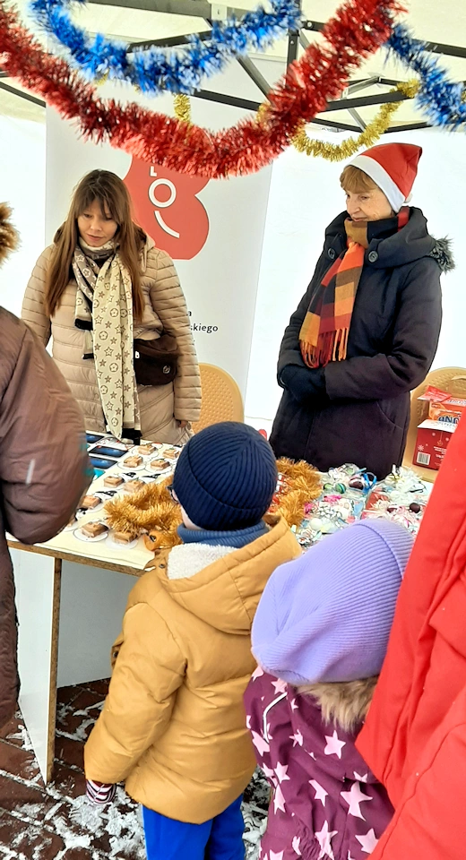 Stoisko biblioteczne na grudniowych stegnaliach. Dwie panie za stoiskiem uśmiecha się do osób naprzeciwko. A tam stoi kobieta w brązowym płaszczu i dwójka dzieci, chłopiec i dziewczynka.