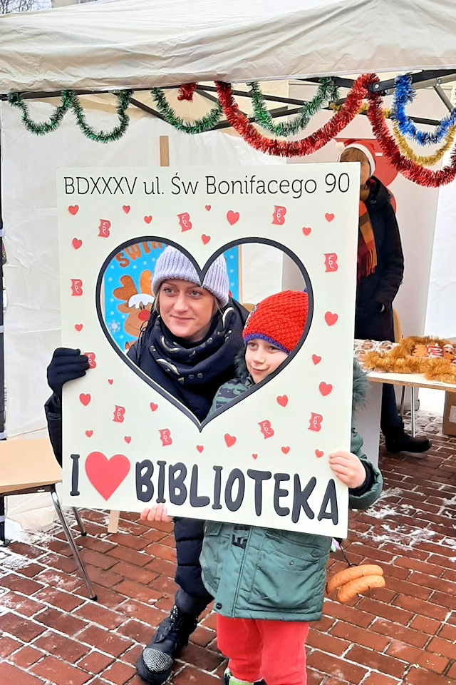 Świąteczna foto ramka, a w niej matka z dzieckiem. W tle stoisko biblioteczne na grudniowych stenaliach.