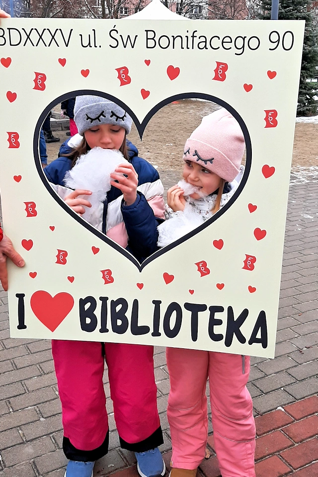 Biblioteczna foto ramka, a w niej dwójka dzieci w zimowych ubraniach jedzą białą watę cukrową. W tle blok i kilka drzew bez liści i jedno drzewo iglaste.