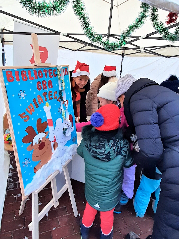 Przed stoiskiem z zainteresowaniem stoi dwójka dzieci i kobieta w zimowych ubraniach. Po ich lewej stoi tablica z plakatem. Na plakacie renifer i miś polarny uśmiechają się, a nad nimi tekst: biblioteczna gra świąteczna. Za stolikiem zimowo ubrane dwie kobiety w czapkach mikołajowych uśmiechają się do dzieci.