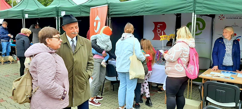 Na chodniku rozłożone są stanowiska promocyjne różnych instytucji, w tym biblioteki mokotowskiej. Koło stanowisk kręcą się całe rodziny. Na pierwszym planie starszy mężczyzna i kobieta obserwują ruch wokół stanowisk.