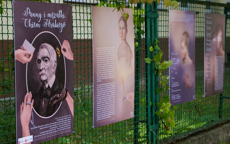 Na drucianym płocie widać cztery powieszone plansze. Na trzech z nich poza tekstem są narysowane młode damy z czasów zaborów. Na czwartej, najbliżej obiektywu, widać portret hrabiego, starszego, siwego mężczyzny.