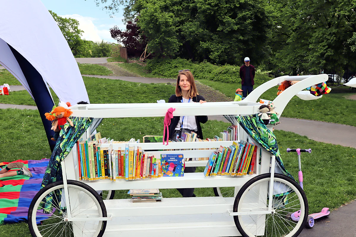 NA tle zielonego parku stoi bardzo duży biały wózek zapełniony książkami dla dzieci i udekorowany kilkoma szmacianymi lalkami. Stoi przy nim kobieta w średnim wieku w okularach wplecionych we włosy.