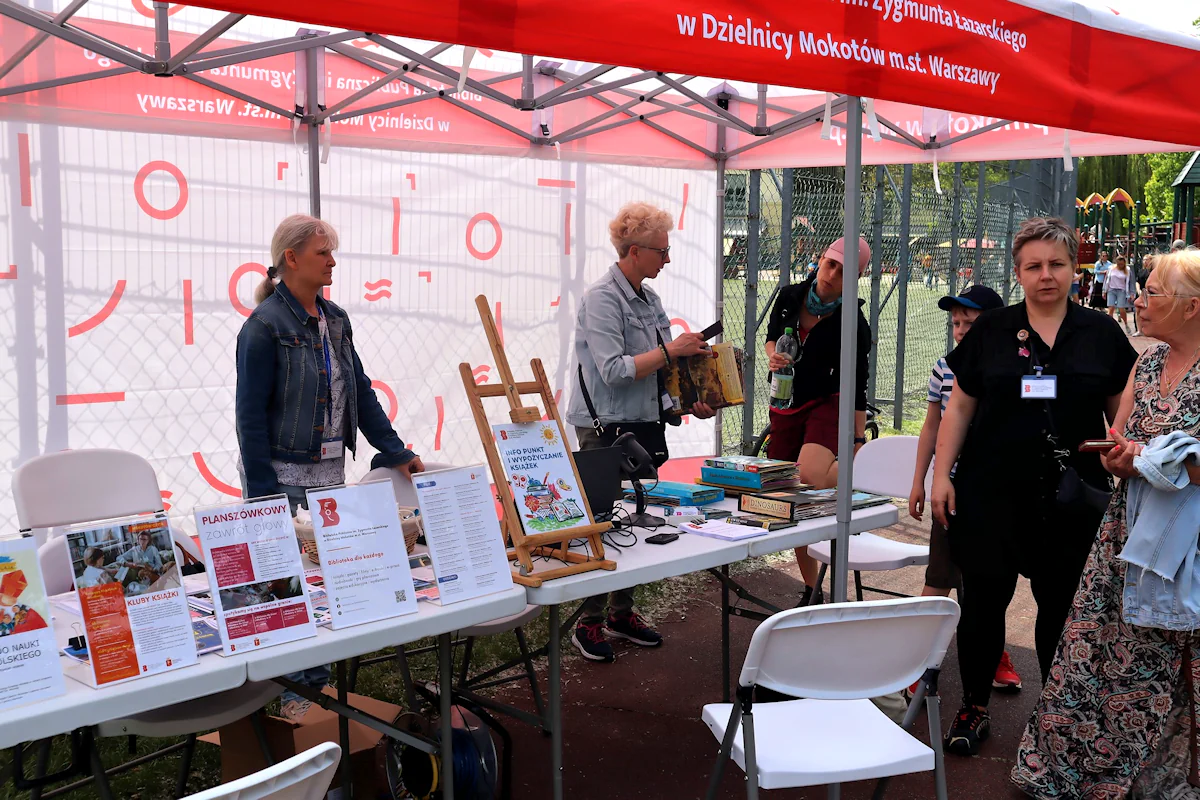 Punkt informacyjny rozłożony na czas pikniku. Pod biało-czerwonym rozkładanym dachem dwa białe rozkładane stoły z ulotkami. Za stołem dwie kobiety w średnim wieku. Jedna z nich rozmawia z młodą kobietą. Dwie inne kobiety rozmawiają  przed stolikami.