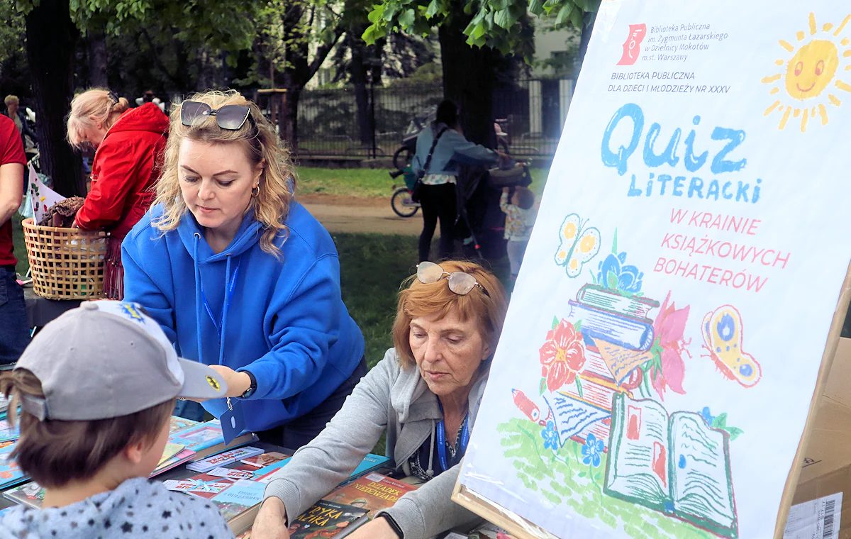 Dwie bibliotekarki układają coś na stole. Dziecko wpatruje się w stół. Z prawej duży plakat z narysowanym słoneczkiem i kilkoma książkami. Na nim napis: quiz literacki w krainie książkowych bohaterów.