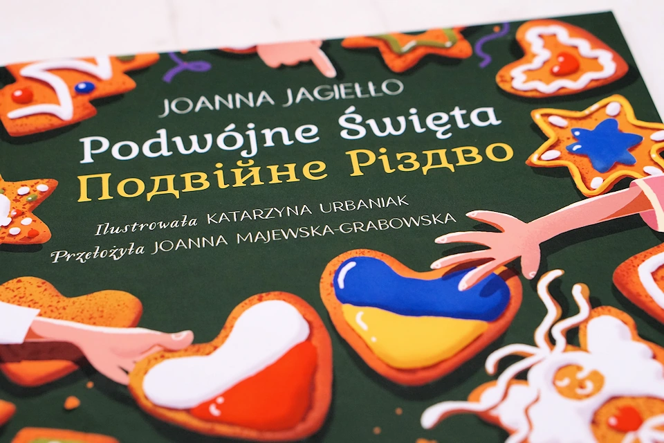 Okładka książki Joanny Jagiełło, Podwójne Święta. Tytuł po polsku i ukraińsku, a pod nim ręce z piernikami w ręku. Piernik są w barwach polskiej i ukraińskiej flagi.