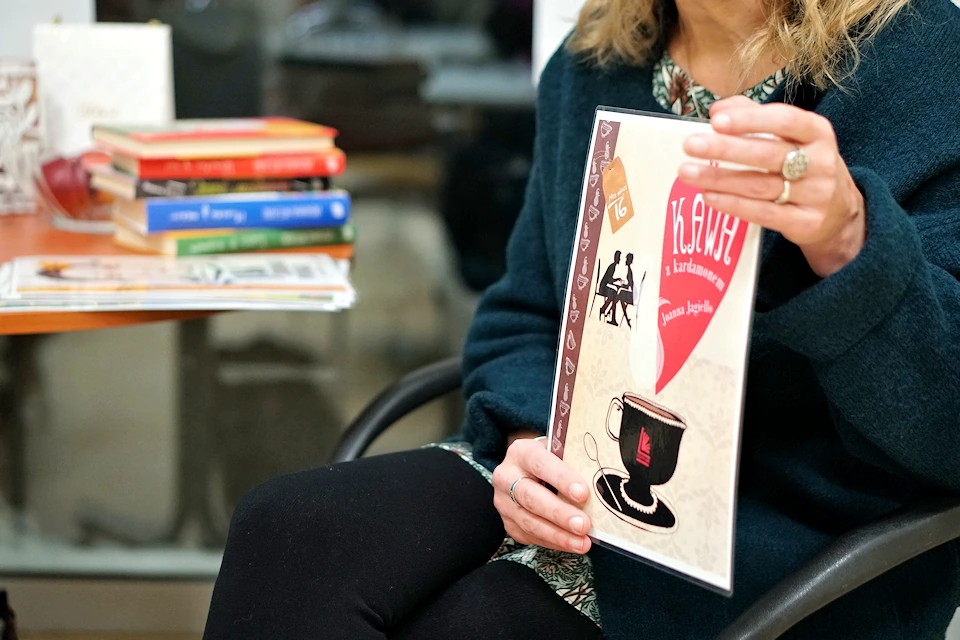 Joanna Jagiełło siedzi na krześle z plakatem książki w ręku. Prezentuje on tytuł Kawa z kardamonem, gdzie obok rysunku filiżanki parującej kawy, gdzieś w głębi siedzi para przy stoliku. Obok autorki na stoliku leży kilka książek.