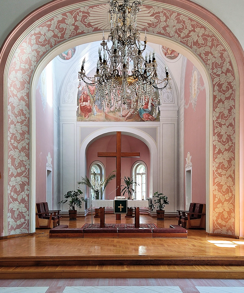 Ołtarz kościoła ewangelicko-augsburskiego. Wnęka ołtarza otoczona ozdobnym motywem białych liści na fioletowym tle. Z góry zwisa trójpoziomowy duży żyrandol. Przed stołem ołtarzowym klęczniki. Stół z białym obrusem. Za stołem drewniany krzyż bez żadnych ozdób. Za krzyżem mniejsza wnęka z dwoma oknami i jasnoróżowymi ścianami. Nad wnęką religijny malunek zasłonięty przez żyrandol.