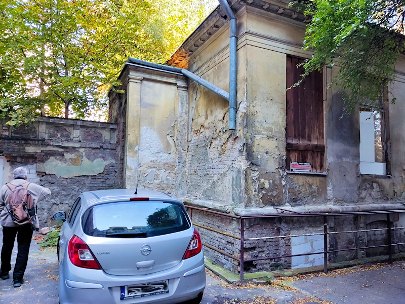 Parterowy budynek w złym stanie. Odrapany tynk, okna zabite dechami. Przed budynkiem parkuje srebrny samochód kompaktowy. Za domem rosną drzewa liściaste.