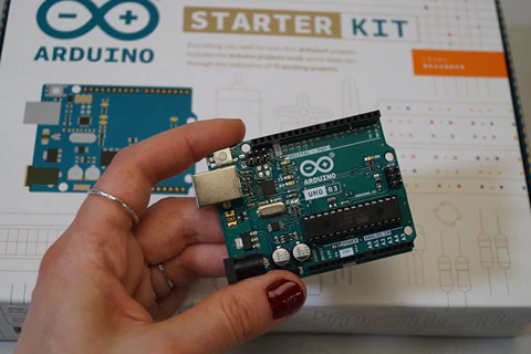 Kobieca ręka trzymająca płytkę Arduino - mikroprocesor. W tle widać opakowanie od zestawu Arduino Starter KIT.