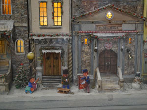 Model miniaturowej kamienicy. Przed zaśnieżoną kamienicą widać figurki dorosłych i dzieci. W oknach kamienicy palą się światła.