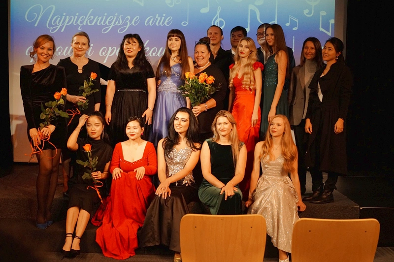 Zdjęcie grupowe studentek i studentów szkoły muzycznej w uroczystych strojach. Pięć dziewczyn siedzi na scenie, reszta stoi za nimi.
