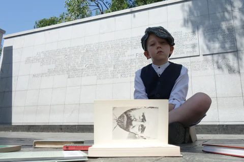 Chłopiec ubrany w stylu przedwojennym siedzi na tle pomnika Umschlagplatz (jansno marmurowa ściana z wyrytym tekstem upamiętniającym ofiary Holokaustu). Przed nim leży książka z widoczną stroną z portretowym zdjęciem Janusza Korczaka.