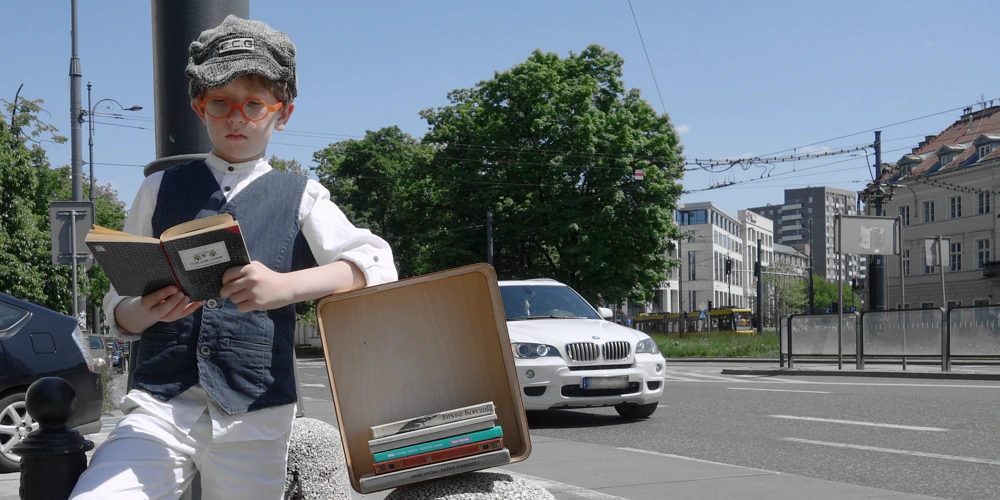 Zdjęcie panoramiczne z widokiem na jezdnię, drzewa, tory tramwajowe i niskie budynki dalej w tle. Na pierwszym planie po lewej chłopiec opiera się o latarnię z książką w ręku, opierając się lewym łokciem o pudełko, w którym widać pięć grzbietów książek Korczaka. Chłopiec jest ubrany styl przedwojenny: białe spodnie, biała koszula, ciemnoszara kamizelka, czapka z daszkiem na głowie i okularami na nosie.