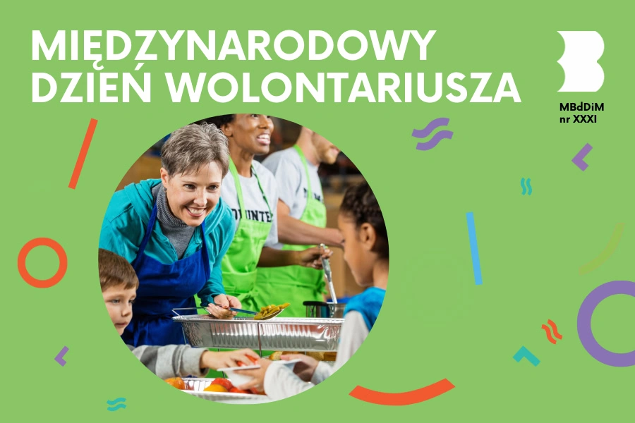 Mały chłopiec kładzie dziewczynce do talerz jabłko. Uśmiecha się do niej kobieta w fartuchu. Dwie inne osoby w zielonych fartuchach stoją przy stole z jedzeniem.
