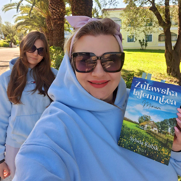 Uśmiechnięta kobieta w niebieskiej bluzie z kapturem, w dużych przeciwsłonecznych okularach trzyma w ręku swoją książkę z serii Żuławska tajemnica.